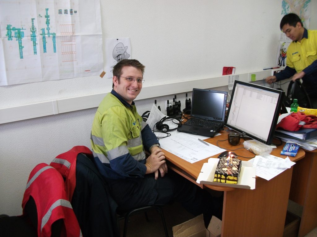 Man sitting at a desk covered in paperwork and working at a computer, looking over his shoulder at the camera.