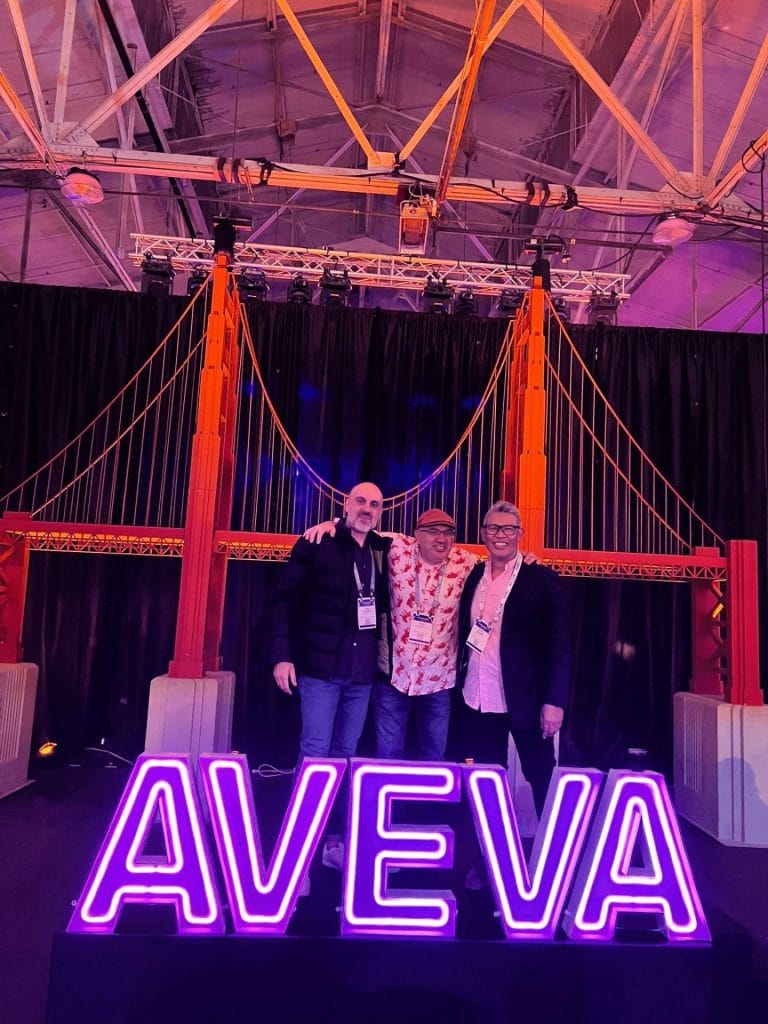 Three men standing behind a lit up, purple AVEVA sign, with a background of a fake Golden Gate Bridge.